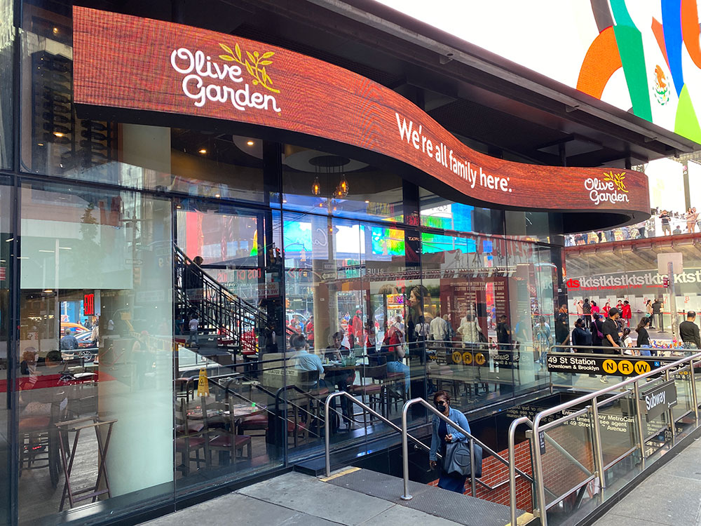 The Olive Garden restaurant in Times Square arrived in New York City just as the theater district was being revived. Here the restaurant sign has been updated to an LED screen and towers over the subway entrance at 49th Street